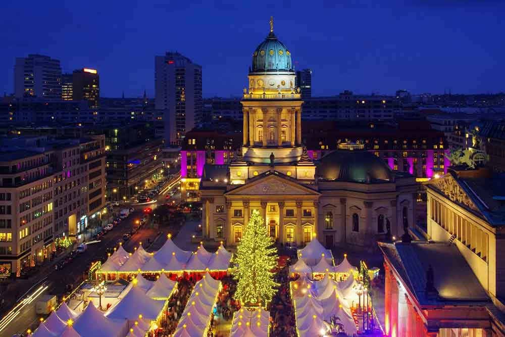 Christmas markets in Belin at night lit up in front of a town hall. There is a chirstmas tree in the centre