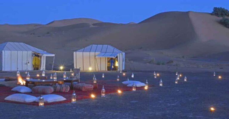 Tow tents in the desert at dusk surrounded by small lanterns. This is the features image for an article about glamping in morocco on a sahara desert camp.