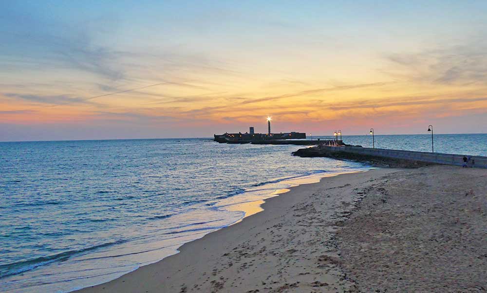 Cadiz beach at sunset with the pier and lighthouse in the background. The lighthouse has a light at the top. The sky is orange and pink the sea is calm. The beach is empty. 