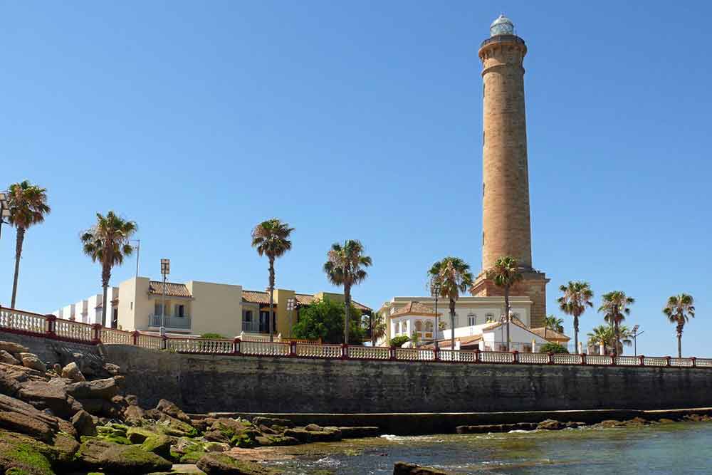 A red bricked lighthouse oat Chipiona Beach stood on a pier with a row of palm trees in front and the sea at low tide.