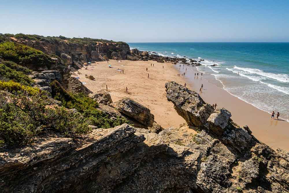 A seculded beach at Coves de Roche with a cove surrounded by rocks, trees and shrubs. There are jagged rocks makeing a crescent shaped bay. 