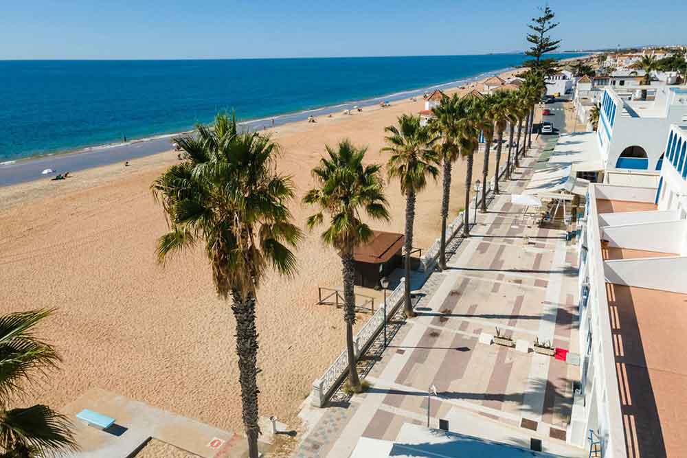 The beach promenade at Islantilla in Huelva with a wide pedestrainised walk way lined with palm trees and shops. The beach is quiet and the sea is brigh blue and calm. 