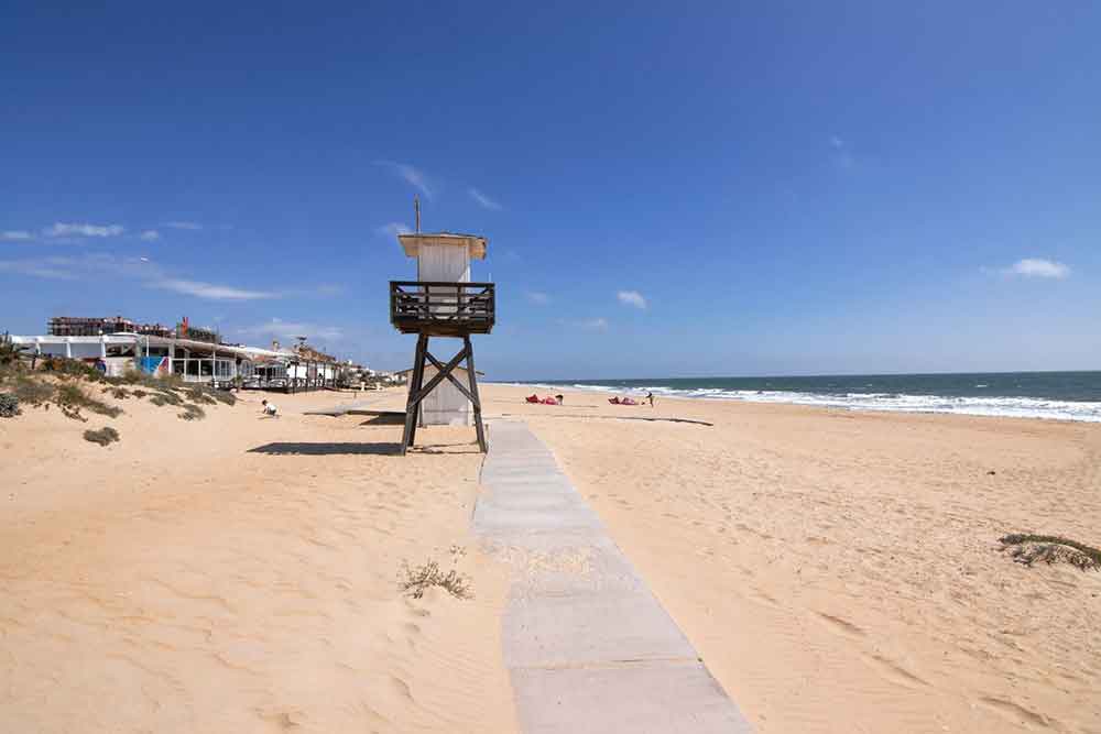 A wide and golden sandy beach at Punta Umbria beach near Seville. There is a boardwalk on the beach and a lifeguard look out booth. There are small shops and vendors at the beach. 