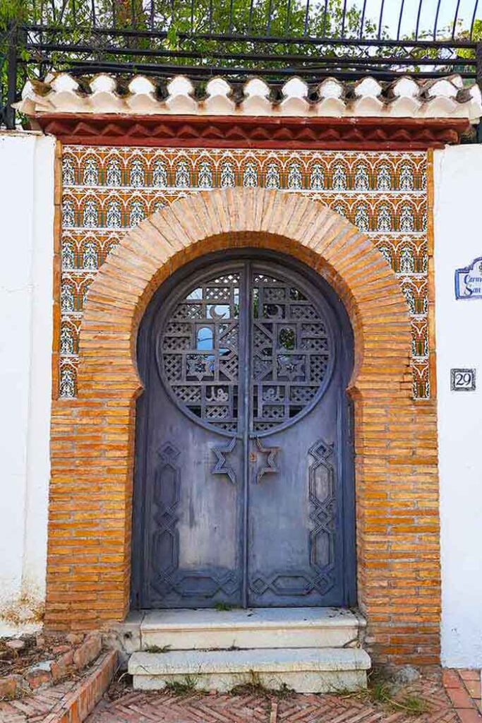 A blacl door with arabic style latticework and decoration on it in the Albaicin area. It's set in a stone brick wall with tile detailing on the upper half. 