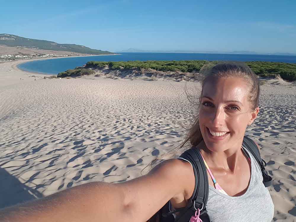 An image of me walking up the sand dunes of Bolonia. There are thoudans of footprints in the the sand, and the dune looks down on a bright blue bay below with grall and hills in the background. 