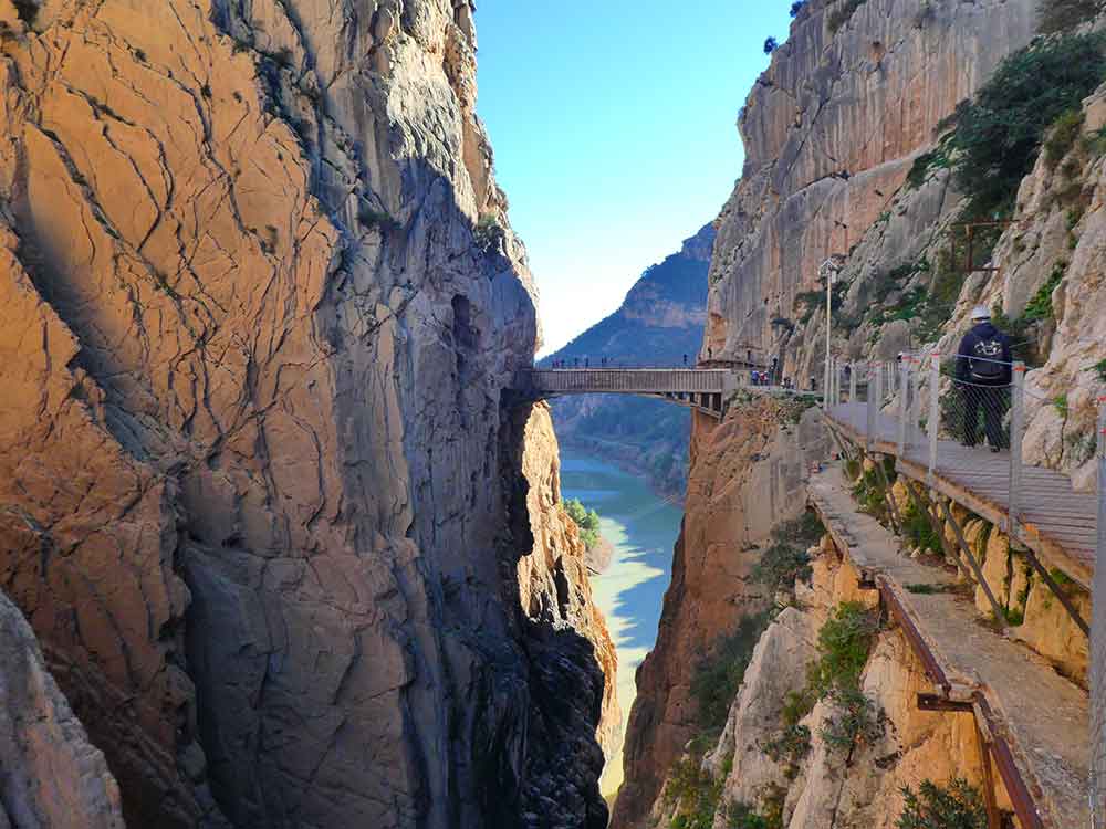 A bridge stretching between two sides of a gorge with a river below and a resivoir in the background. There is a small boardwalk along the left side of the cliff with the remains of the old walkway under it. 