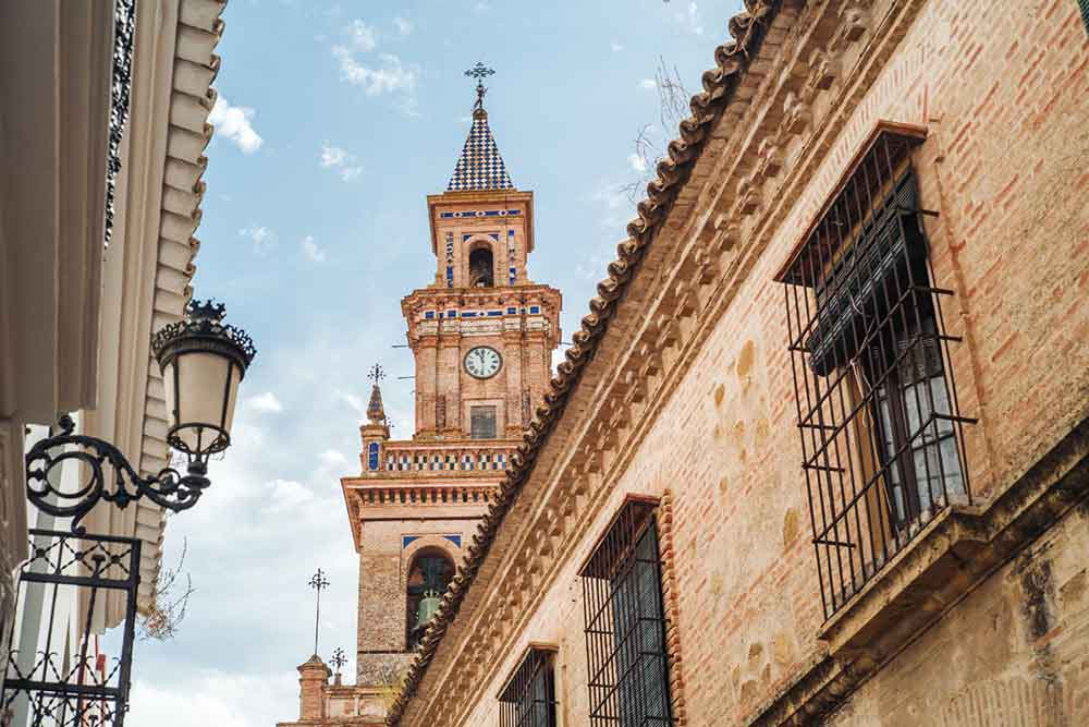 Looking up in the streets of Carmona with old buildings with gates in front of the windows and a lantern on one wall. There is a tower in the background in reddish bricks and decorative white and blue tiles. 