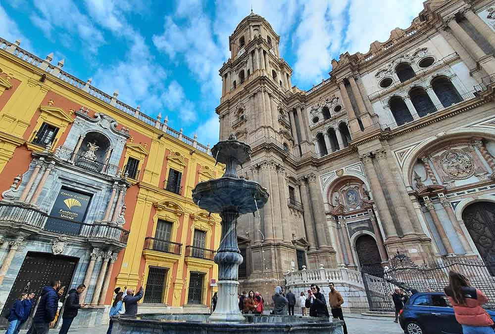 Malaga cathedral with decorative fascade with arched windows and motifs over the arched doorways. Next to it is a red and yellow building with a grey sculpted entrance way. In the forground is an ornate fountain with two tiers.