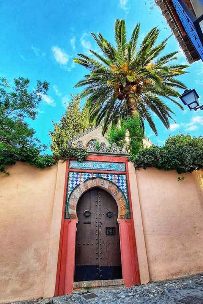 A door in the Albaicin district with colourful arabic decoration. The surround is red with decorative colourful tiles on top The wooden door has a pointed top. There is a palm tree behind it.