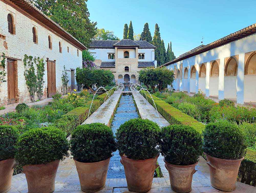 A patio inside the Generalife surrounded by buildings. The buldings are a mix of brick and white plaster with decorative arches. The patio has a long rectangualr pond in the centre with small arched fountains shooting into it. The patio is decorated with bushes, shrubs, flowers and trees.