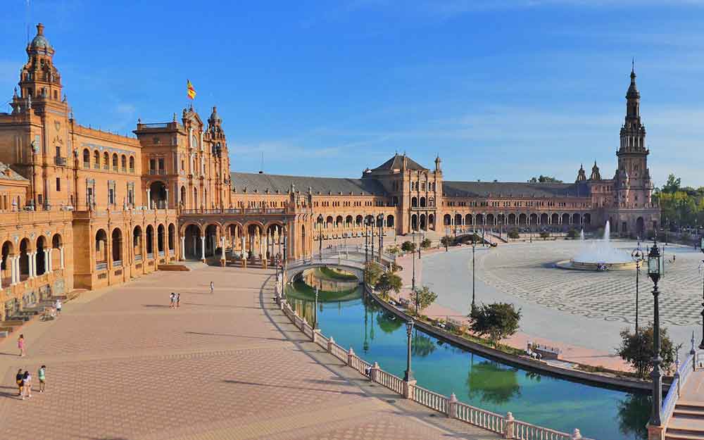 The crescent shaped building of Plaza de España with red bricked patio and orange bricked building. The building is ornate with alcoves and tiles. There is a moat running parallel with bridges crossing it and a fountain the centre of the semi-circle square.