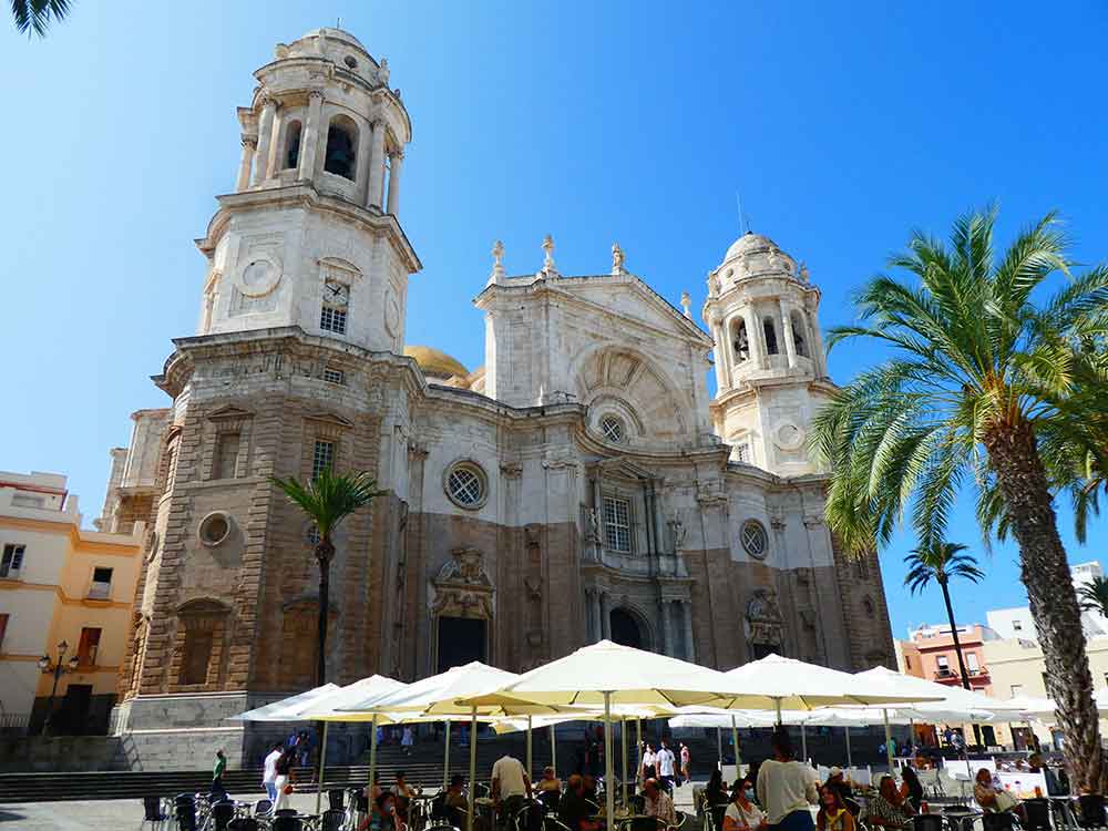 The fascade of Cadiz cathedral with two large towers and a giant central tower. The upper half is pale stone, the lower half is brown stone. In front is a plaza with umbreallas and people drinking at a cafe.
