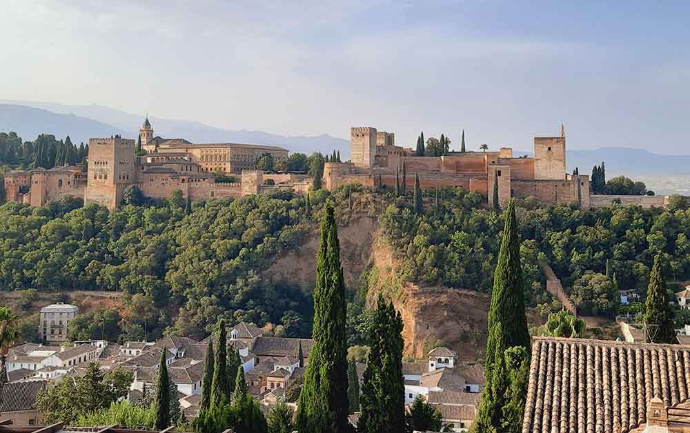 The Alhambra on a hill surrounded by trees and the Albaicn district. The view of the Sierra Nevada mountains can be seen in the background. 