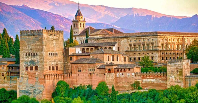 The Alhambra palace in Granada sat on a hill with mountains in teh background and forest in the foreground. Visit as a part of your Spain and Portugal itinerary.