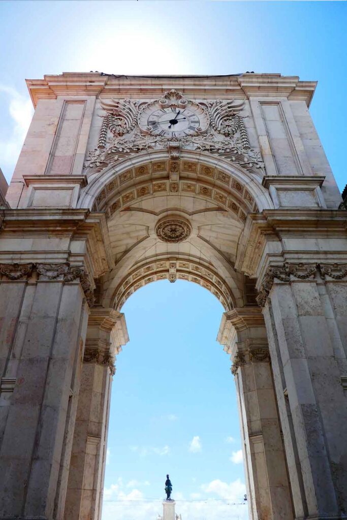 Looking up at the Arco de Rue Augusta. The triumphal arch has an ornate detailat the top with flowers, a shell and wings. There is a clock int he middle. 