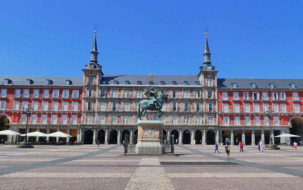 Plaza Major in Madrid, Spain. A big open plaza with a stature in the centre. The statue is of a hourse and a knight and is on a plinth, there is a small gate around it. In the background you can see buldings that surround the Plaza. They are red and pale orange. Some of the buildings have spires. There is a small archway that's on the bottom level of the buildings leading around the plaza. Be sure to visit Plaza major on your Madrid 2 day Itinerary or as part of your Portugal and Spain 14 days itinerary