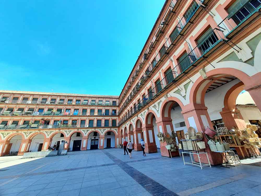 The Best Cordoba in One Day Itinerary 16 Plaza de la Corredera surrounded by buildings. There are three levels with windows and balconies, Under these levels is a arched arcade with shops.