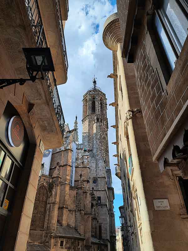 Looking up through the narrow streets of the Gothic Quarter in Barelona. There are ornate buildings in different styles, with decorative turrets, gargoyals and columns on them. 