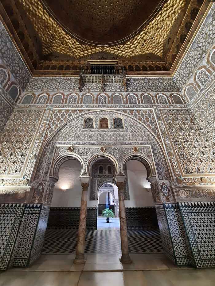 The Salón de Embajadores at the Real Alcazar a square room with ornate platsterwork on the walls and domed gold ceiling. There are pillars with arabic shaped door ways leading through to another hall.