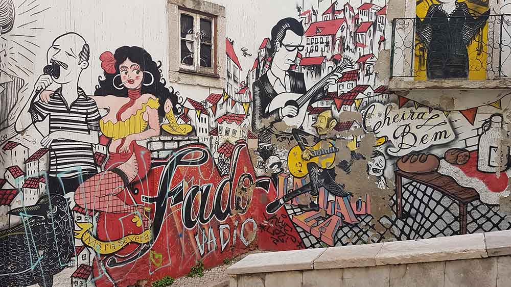 A large mural on a wall in Lisbons Alfama disrict with the words Fado written on it. It's surrounded by cartoon like images of people and scenes of Lisbon. 