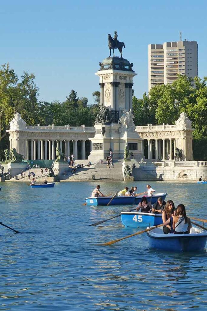 People in blue rowing boats in parque we Retiro in Madrid. There is a large monyment in the background with a lower building with coloumns and a taller column with a horse on top. 