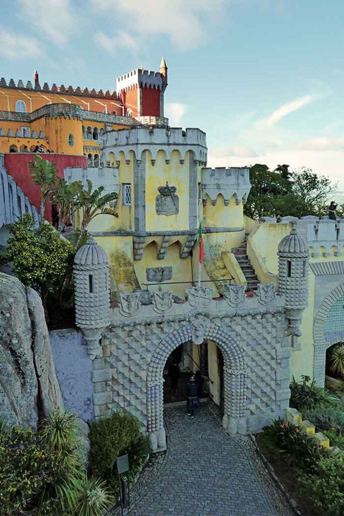 Entrance to Palacio da Pena with a mix of architecural styles and colours. In front is a grey spikey arched gateway. Behind is a light yellow turreted builidng. Behind that are yellow and red turreted buildings. 