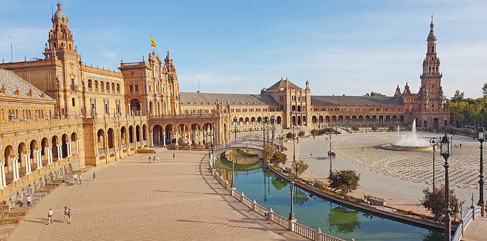 Plaza de España in Seville. The crescent shaped reddish bricked building with towers, balconies, and arcaded walkway. There is a moat running parallel with bridges over it and a fountain in the centre. 