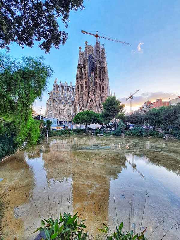 view of Sagrada Familia from across a lake. The building is reflected inthe lake. There is a crane and scafolding on the roof which is still under construction. 
