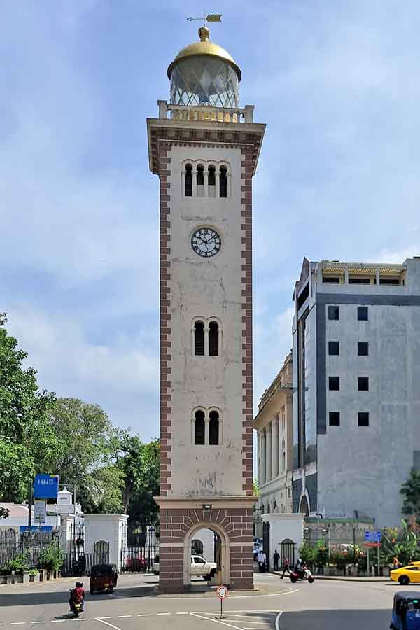 Colombo Fort Old Lighthouse & Clock Tower a rectangular building with an archway through the bottom. There are windows in the walls and a clock. At the top is the glass dome of the lighthouse with a weather vane on top.
