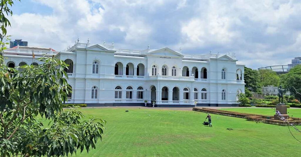 Outside view of the classic style white Colombo National Museum building with the large lawn in front and a man mowing the lawn