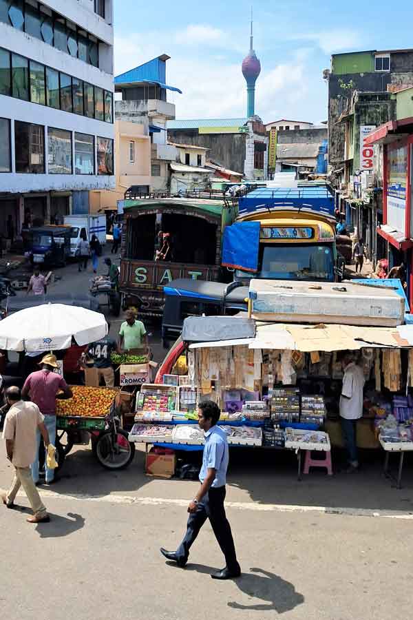 Streets in Pettah district with vendors selling various items such as oranges, and husehold goods. In thebackground is the Lotus Tower. In the foreground are people walking. 