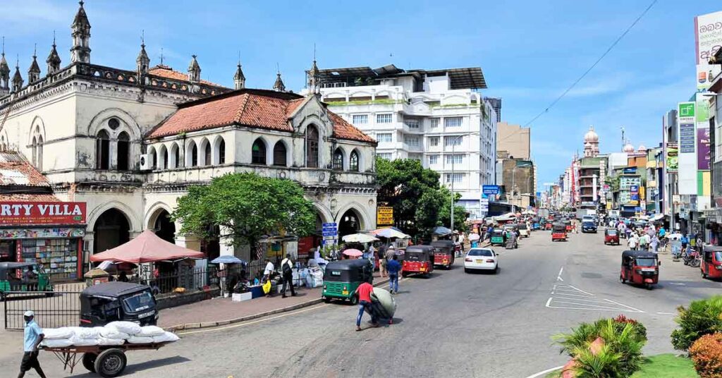 The colonial building of the old town hall. with decorative arches, a red rood and arched entrances all around. In the street there are vendors and tuktuks.  