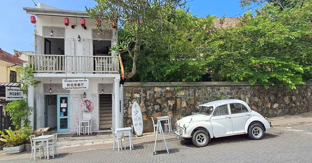 A two store white building with a balcony and red chinese lanterns. The downstairs has a blue door. There are white tables out the front and a white Beetle car. It's on a hill, so the chairs and car are at a slant. 