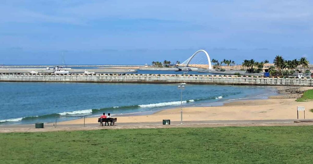 Galle Face Beach promneade with a small sandy beach in the background and waver breakers for the port and an arched shaped bridge in the distance. There are people sat on the bench in the forground.