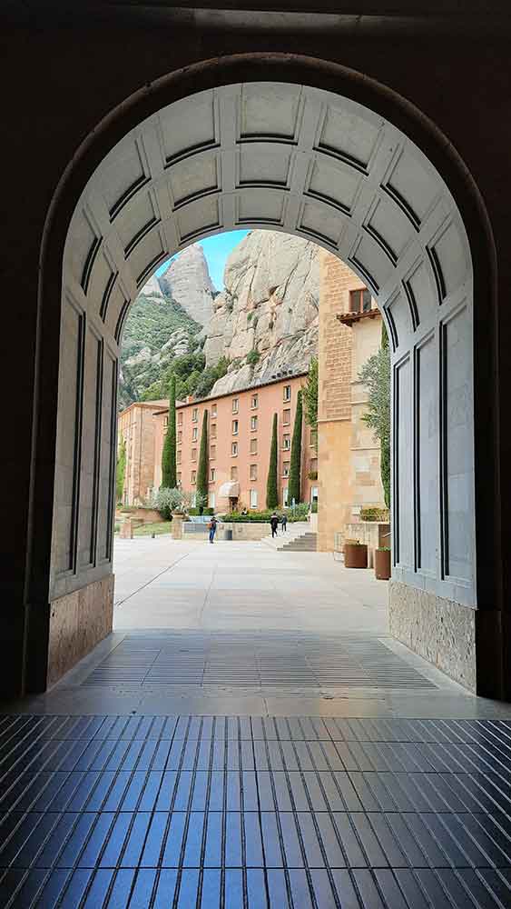 Best Montserrat Tour from Barcelona (With Tapas + Winery Visit) 10 A view looking out through the entrance of the Monastery with buildings and mountains in the background.