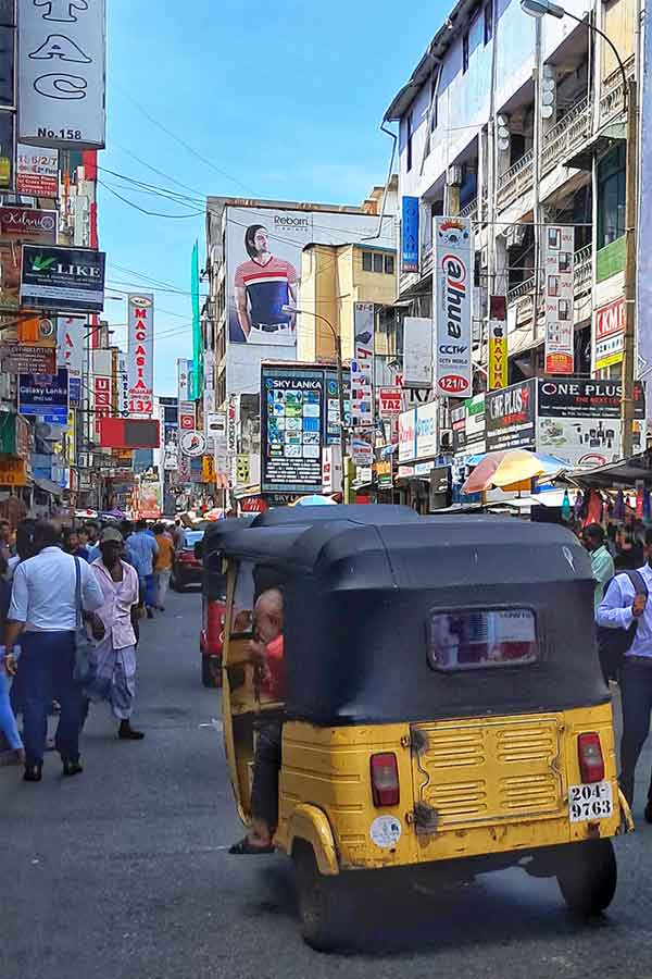 The main high street in Pettah District with signs covering the building walls. There are a lot of people walking and shopping and a yellow tuk-tuk inthe foregound. 