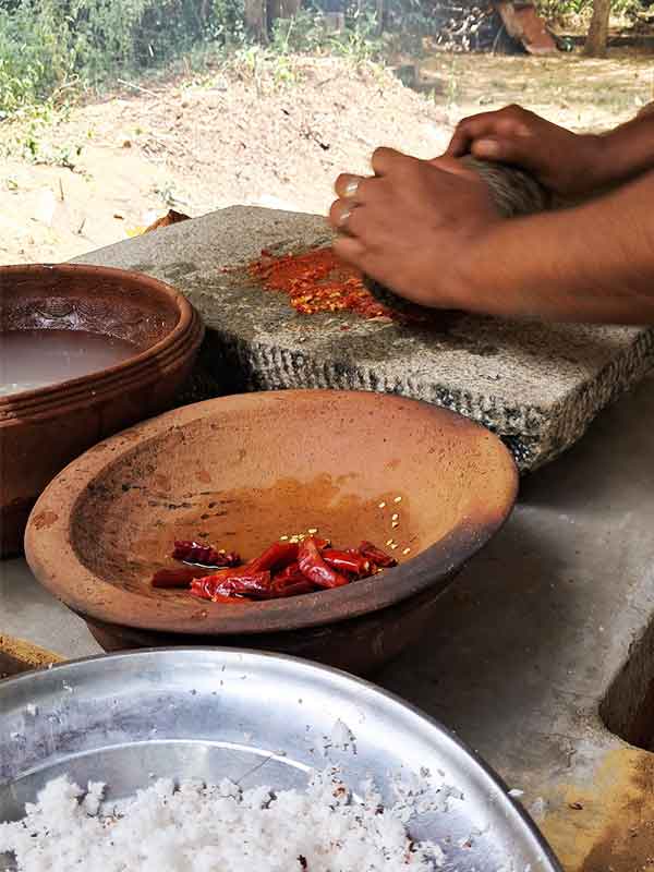 bowls of fresh ingredients to make sambal - there is coconut, chilli and water. A lady is crushing chilis with a stone by hand. 