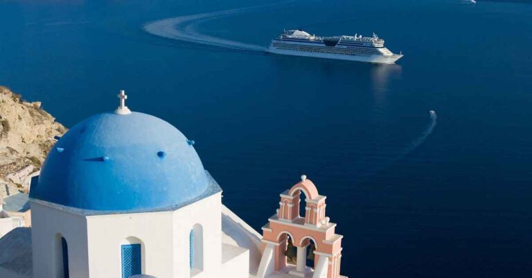 Image of a cruise ship in a calm ocean with a view looking out over a white building with a blue domed roof in Santorini, Greece. This is the featured image about the reasons to take a cruise.