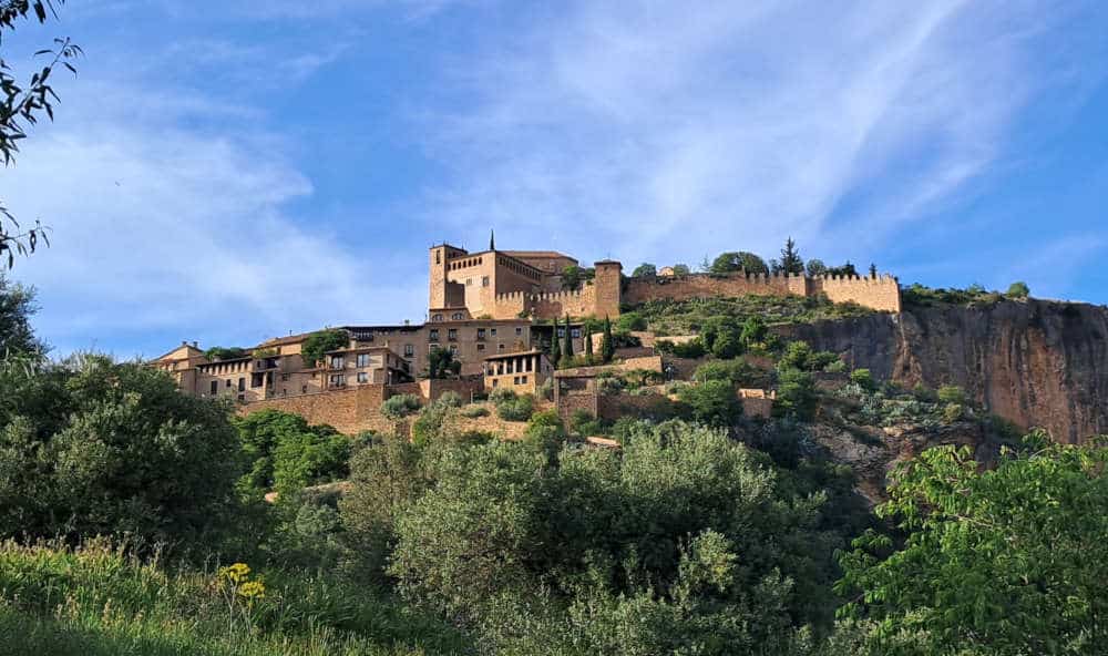 Looking up at Alquézar town from the walkway with the sun hitting the yellow coloured buldings on the rock. There are trees and bushes all around and a blue sky.