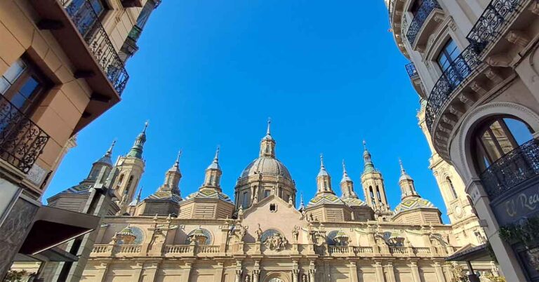 El Pilar Cathedral in Zaragoza with a focus on the decorative roof domes adn spires. There are two buildings in the forground with decorative ralings in front of the windows.