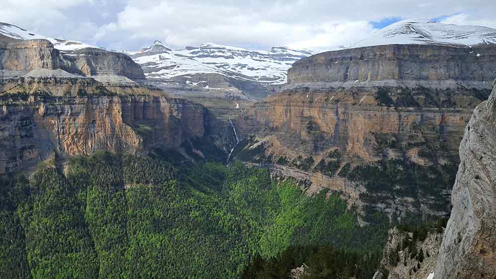 Landscape of Ordesa y Monte Perdido National Park with snow capped mountains in the background, and straight faced mountains in the forground. There are trees in the valley below. A visit to this UNESCO heritage site is one of the best things to see in Aragon.