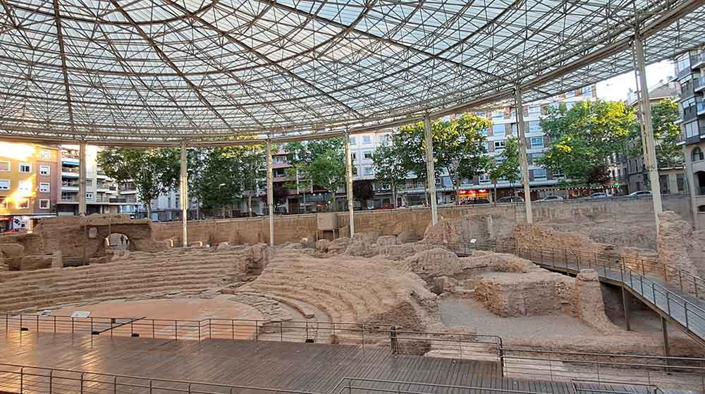 The excavations of the Roman Theatre in Zaragoza with several ros of seats under a large canopy surrounded by the modern city.