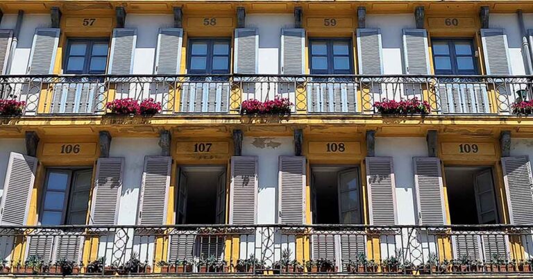 Doors with grey shutters on and numbers above the doors. There is a balcony in front with pink flowers hanging from it. The walls are white and yellow. See these at the Plaza de la Constitución, San Sebastian Old Town. Discover more on is San Sebastian worth visiting in this article.