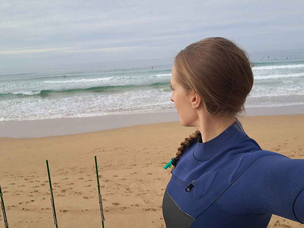 A woman (me) wearing a blue wetsuit looking out at the waves ready to go surfing.