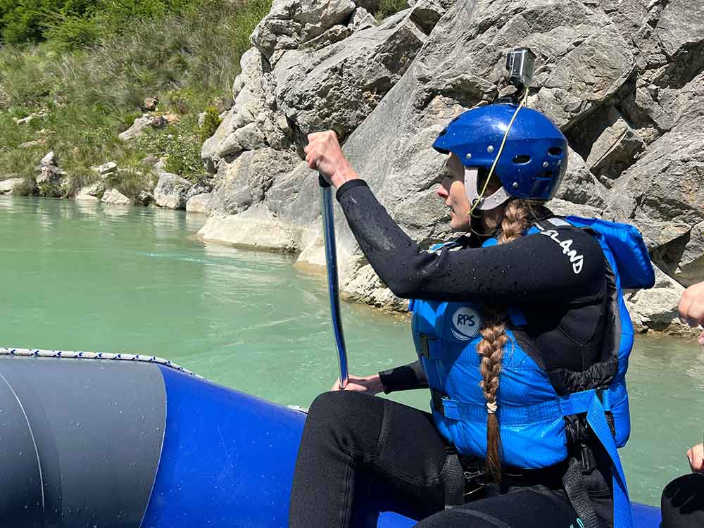 Me holding a paddle, sat in a white water raft. I'm wearing a black wetsuit, blue life jacket and blue helmet.