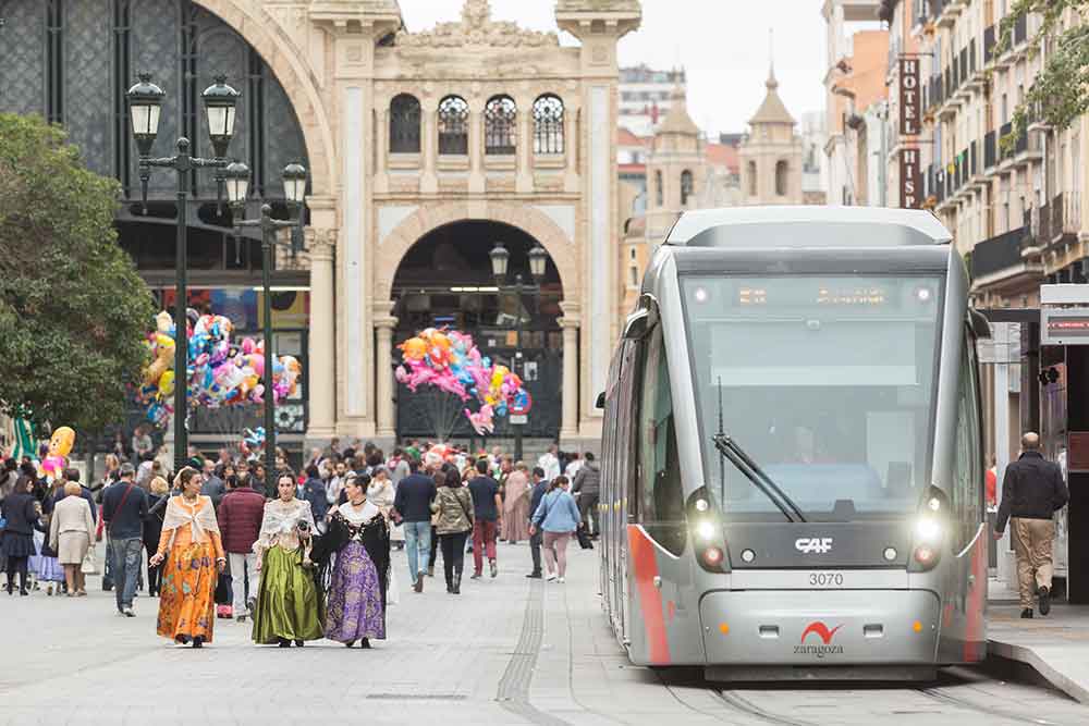 Zaragoza city tram in the centre with the Central Market in the background. There are people in walking in the streets and three ladies wearing traditional costumes.