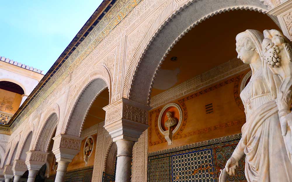 Inside the main courtyard of Casa de Pilatos with decorative arches and a white marble statue of a woman in the front. The back wall has a bust sculpture and decorative tiles. 