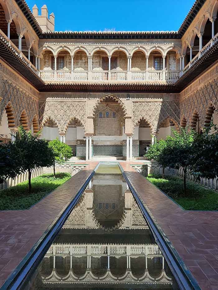 Patio de las Doncellas (The Maidens’ Courtyard) at the Royal Alcazar in Seville. A long recangular pond in a patio surrounded by small shrubs and trees and ornately decorated arcaded walkways.