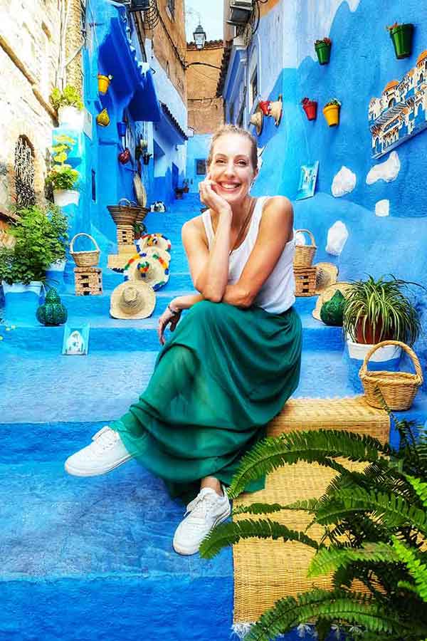 A woman (me) wearing a white sleeveless top and a long green skirt is sitting on the blue street in Chefchaouen - this maxi skirt came to just above my ankles and was the perfect length to wear in Morocco.