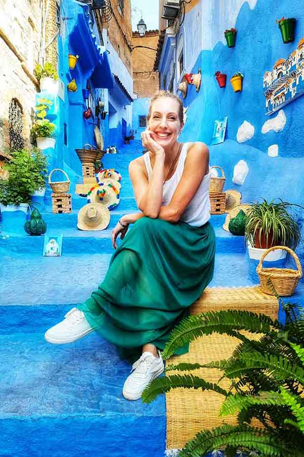 Chefchaouen is known as the blue city, this photo shows blue stairs with lots of potted plants and woven items on it. In the middle is a woman wearing a white vest and green long skirt with white trainers. self-guided 10 days Morocco itinerary from Casablanca to Marrakech